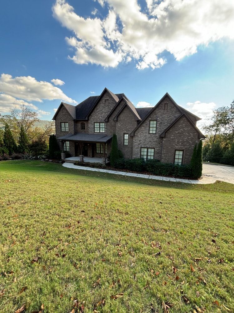 Large brick house with multiple gables on a grassy lawn under a partly cloudy blue sky.