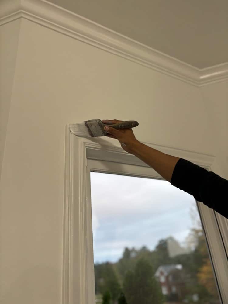 Person painting white trim around a window with a paintbrush, indoors.