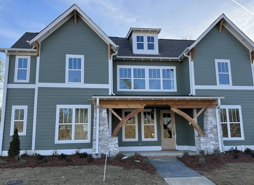 Two-story green house with white trim, stone accents, and wooden porch, under blue sky.