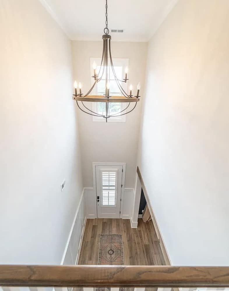 Looking down a long hallway: chandelier, white walls, door with window, wood floors.