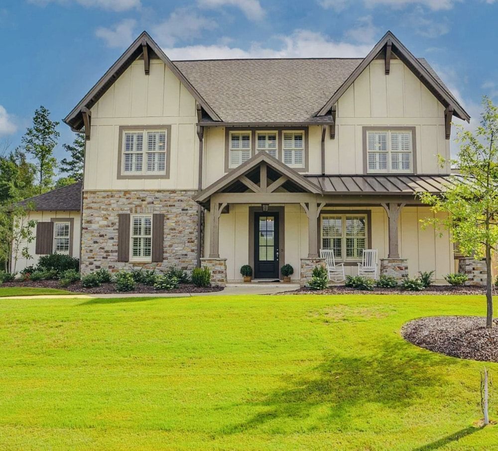 Two-story house with beige siding, stone accents, and a dark gray roof, set on a green lawn.