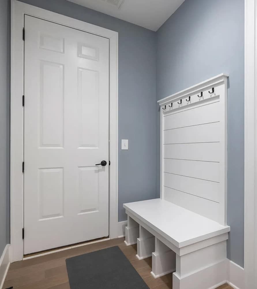 White entryway bench with hooks and door in a room with blue walls and wooden floor.