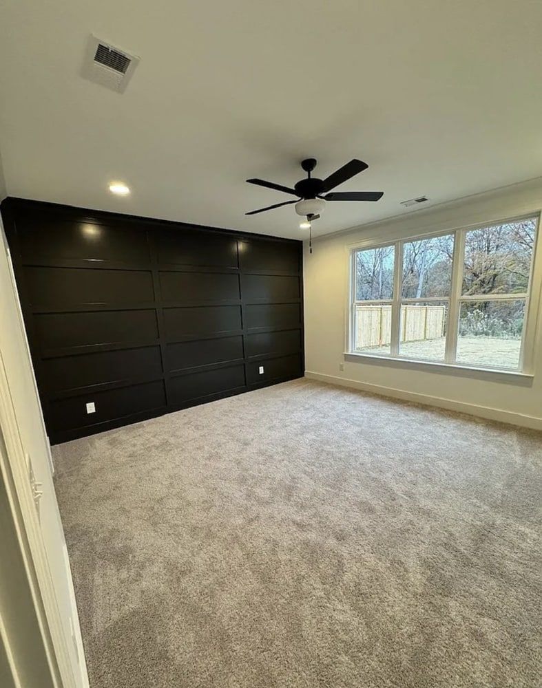 Empty bedroom with black accent wall, grey carpet, and a ceiling fan. Large window with outdoor view.