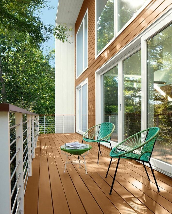 Wooden deck with green chairs, small table, and large windows overlooking trees.