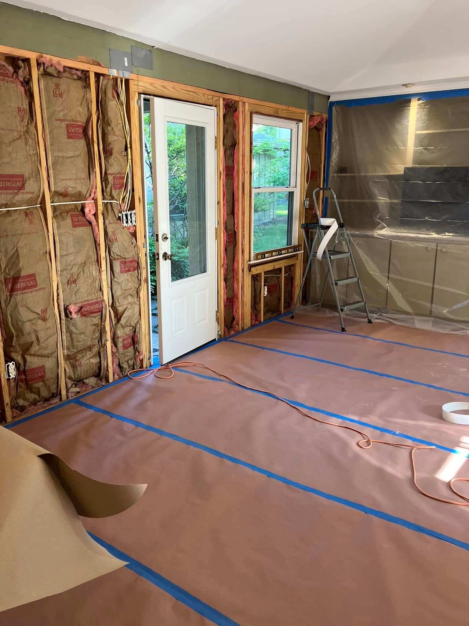 Interior room renovation in progress. Open door, window, exposed insulation, brown floor covering, blue tape.