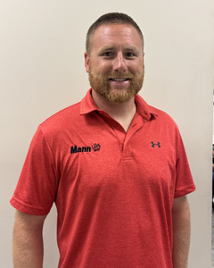 Man in red polo shirt, Mann logo, smiling, standing in front of light wall.