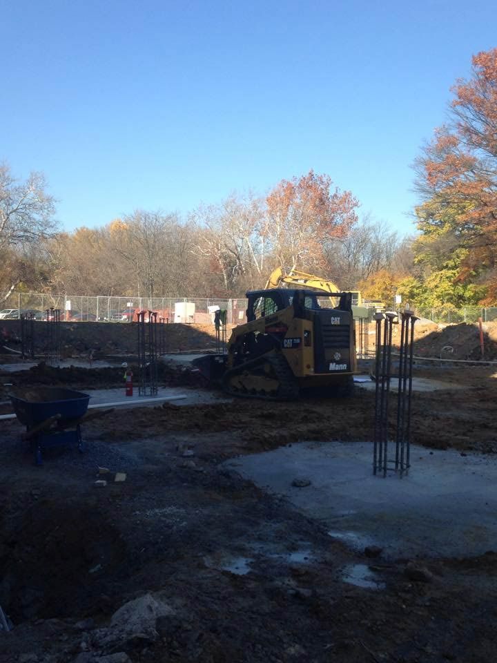Construction site with a skid steer loader on a concrete foundation, trees in the background under a blue sky.