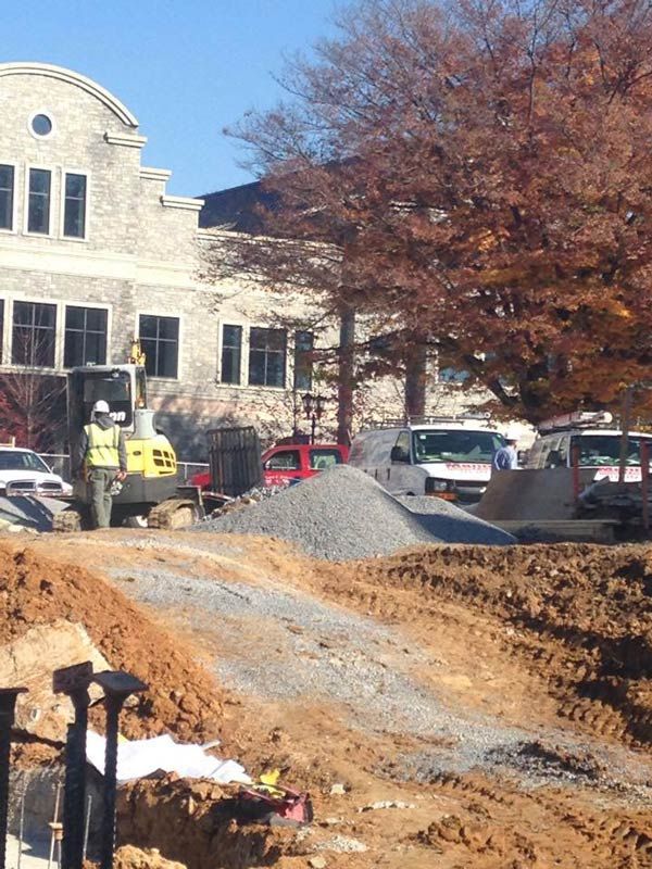 Construction site with an excavator, pile of gravel, and brick building in background.