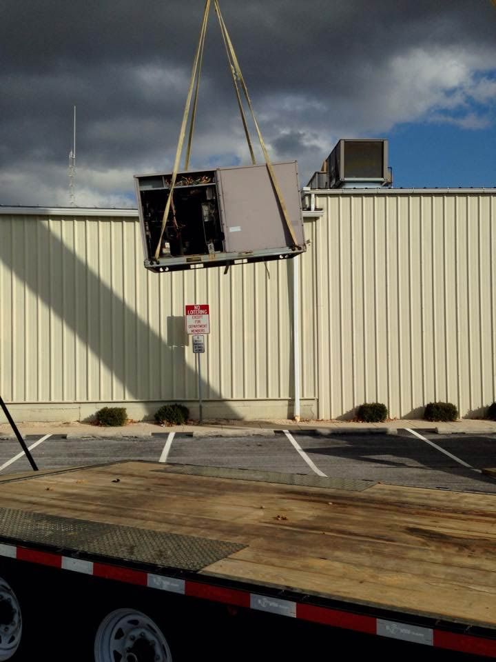HVAC unit suspended by crane over a flatbed truck near a building with a parking lot.