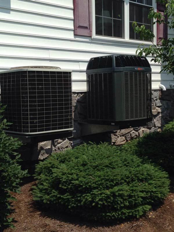 Two air conditioning units sit on a stone wall next to a house with a white exterior.