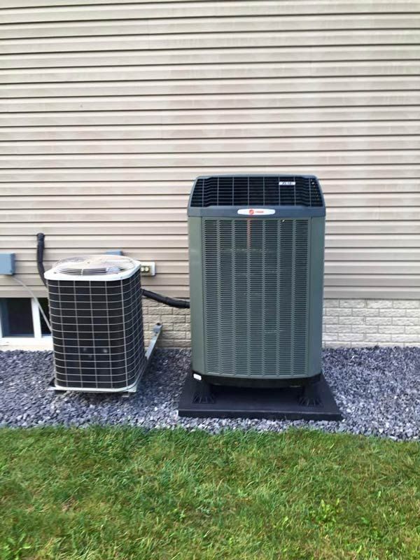 Two air conditioning units side-by-side, outside a building with tan siding and a gravel base, on a grassy lawn.