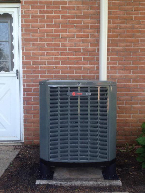 Gray air conditioning unit outside a brick building. White pipe and door present.