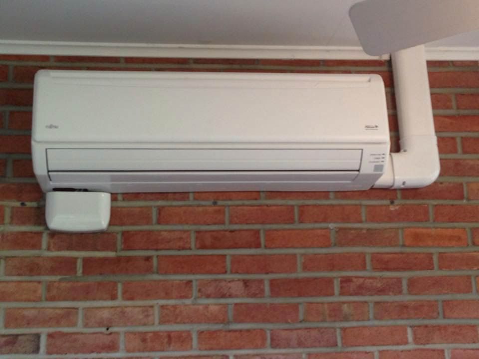 White air conditioning unit mounted on a red brick wall, with a white vent pipe.