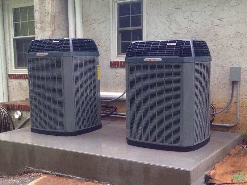 Two grey air conditioning units on a concrete pad next to a building with windows.
