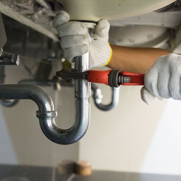 Plumber wearing gloves uses a wrench to work on pipes under a sink.