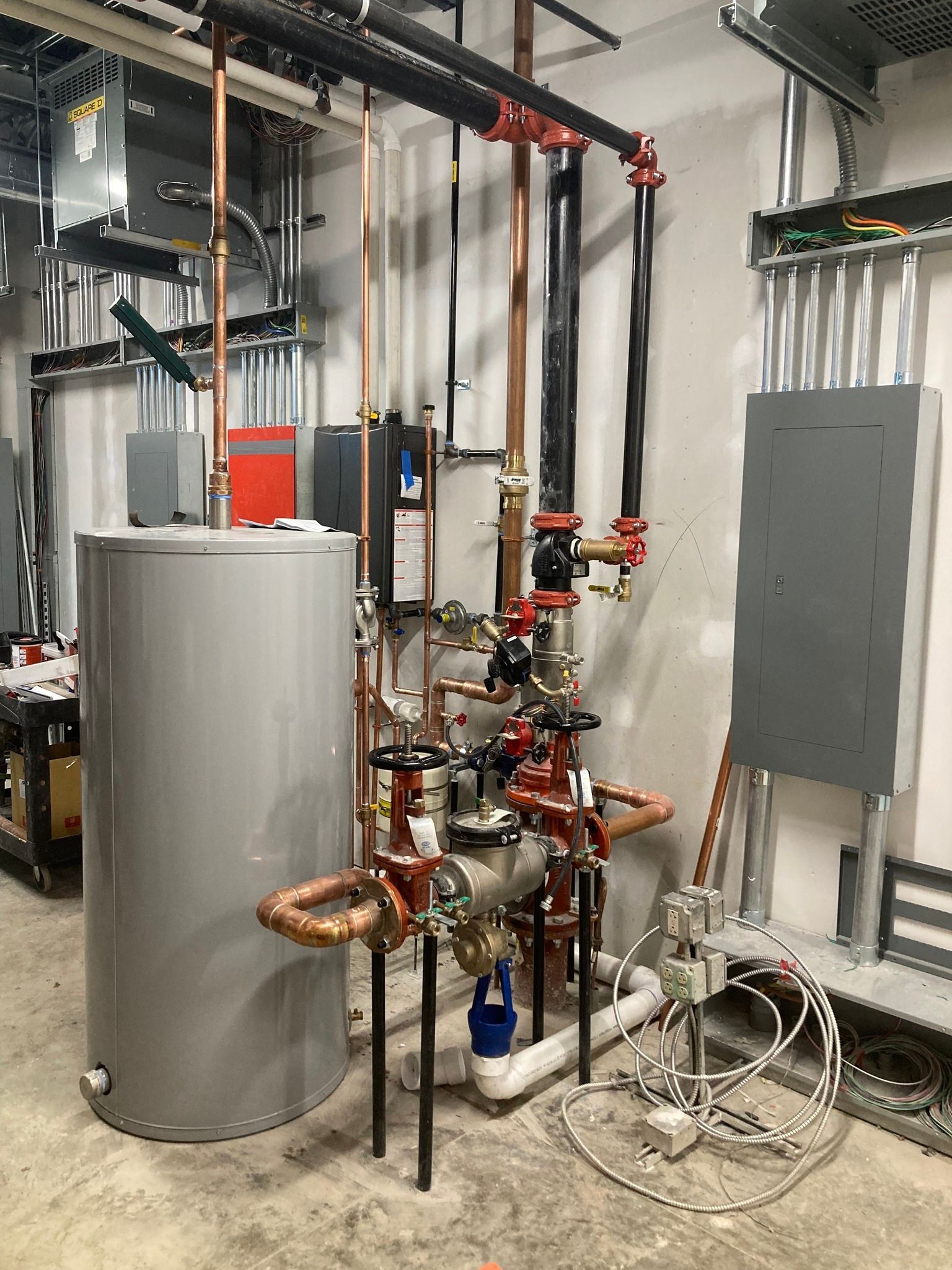 A complex system of pipes, valves, and a water heater in a utility room. Copper and black pipes with gray tank.
