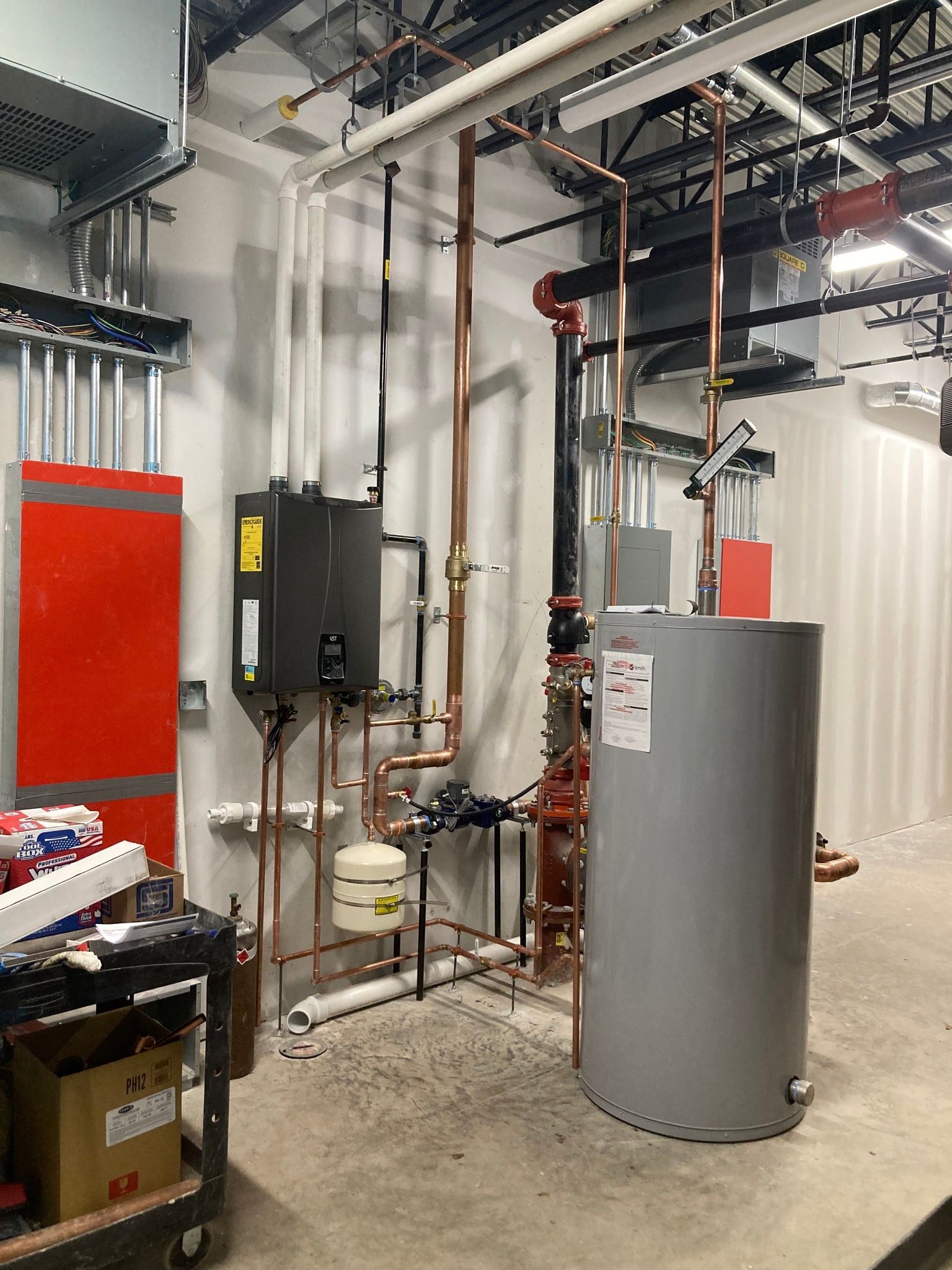 Plumbing and water heater installation against a white wall in an industrial setting. Copper pipes and black water heater.