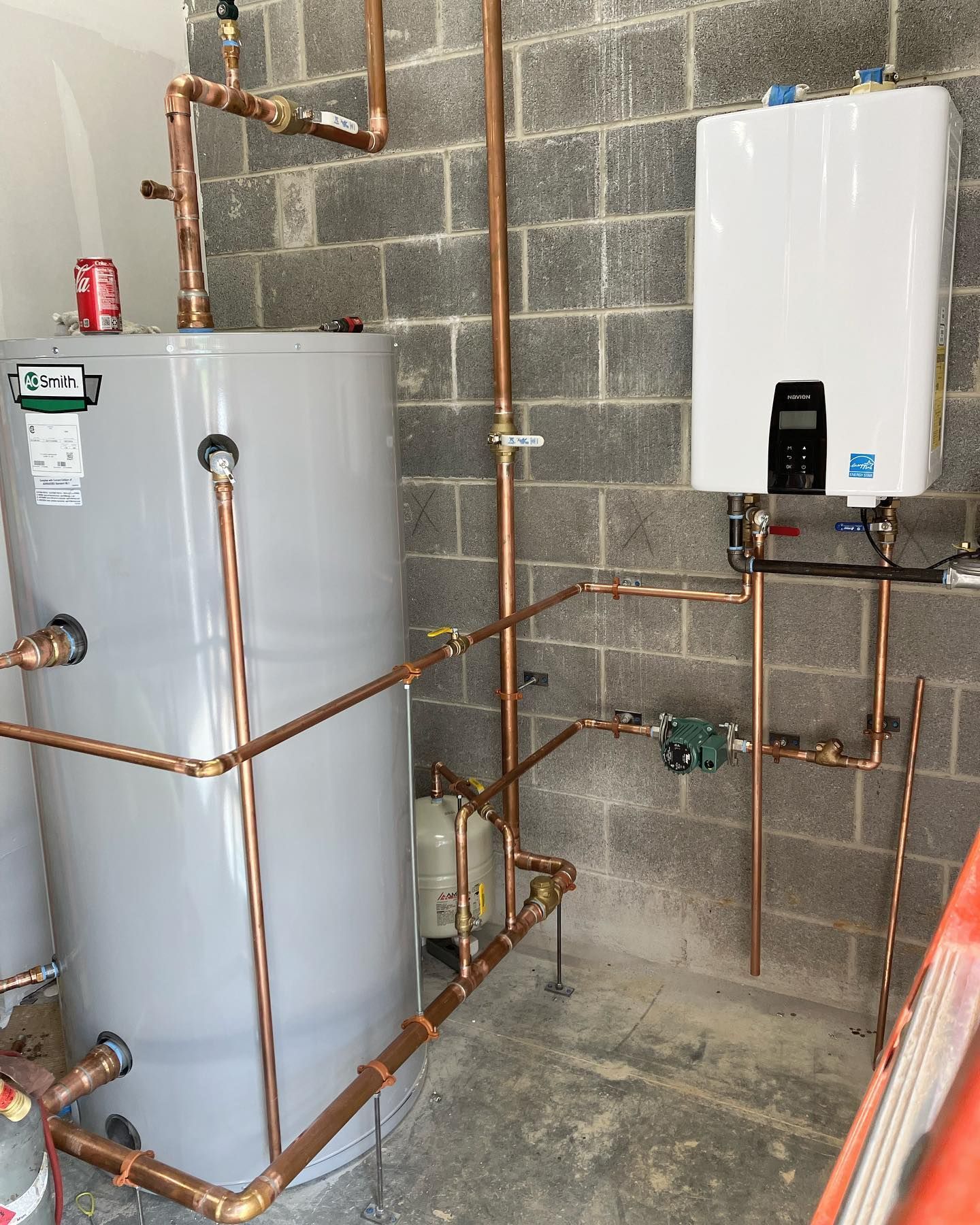 Copper pipes and water heaters against a gray cinder block wall. One is a tank, the other tankless.