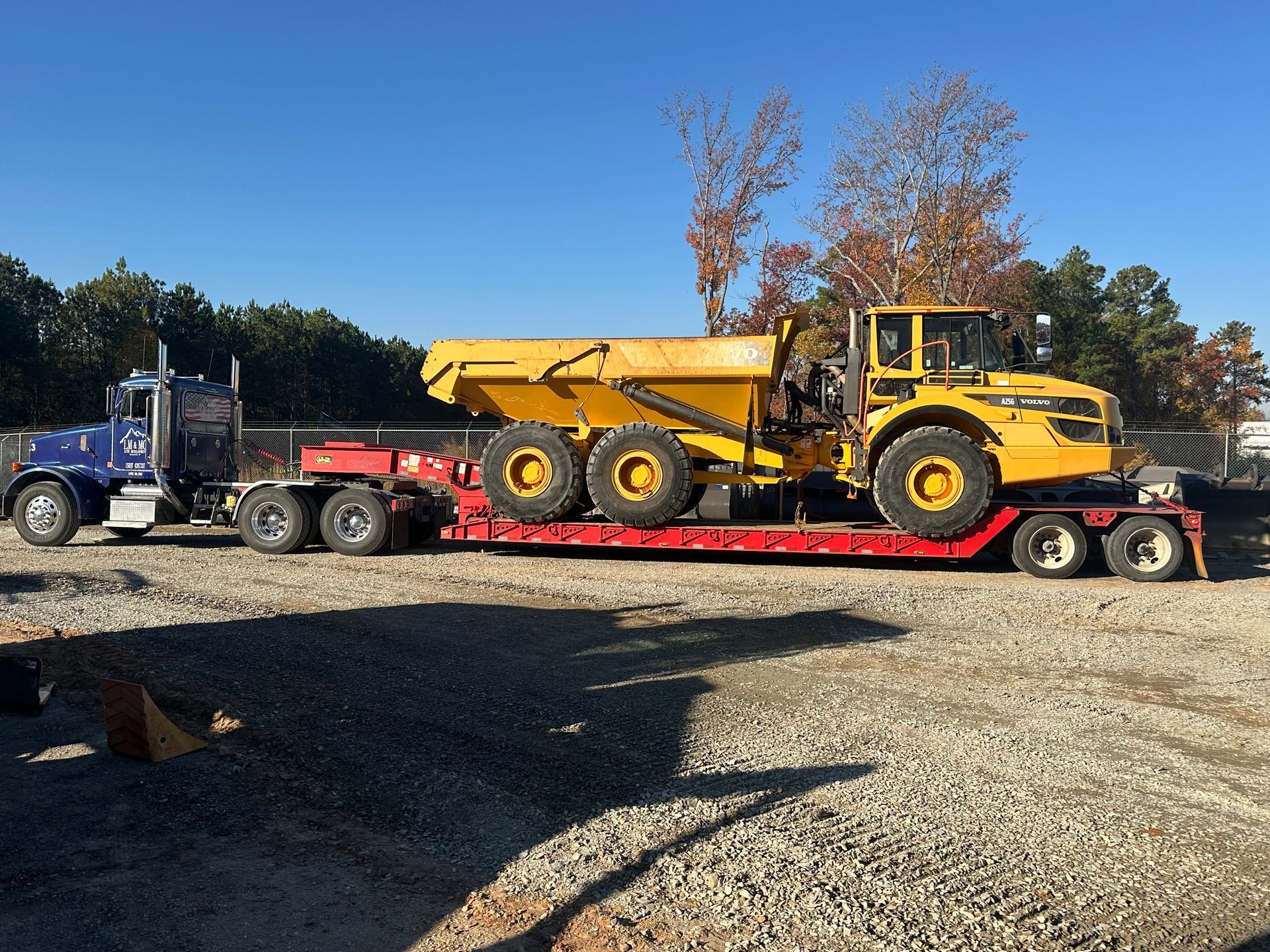 Yellow dump truck on a red lowboy trailer being hauled by a blue semi on a gravel lot under a clear sky