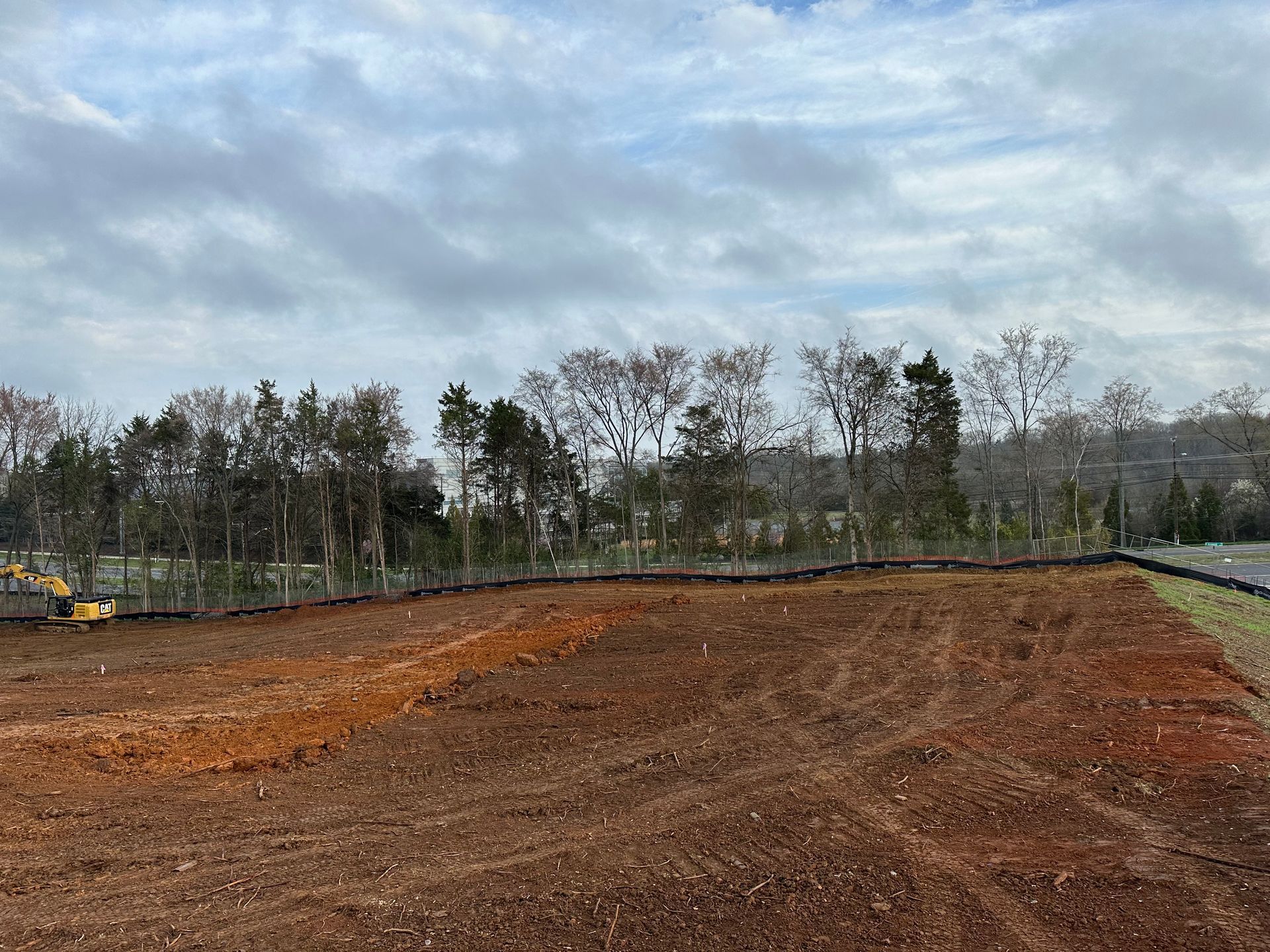 Cleared red dirt lot under cloudy sky, with trees and construction equipment in the distance