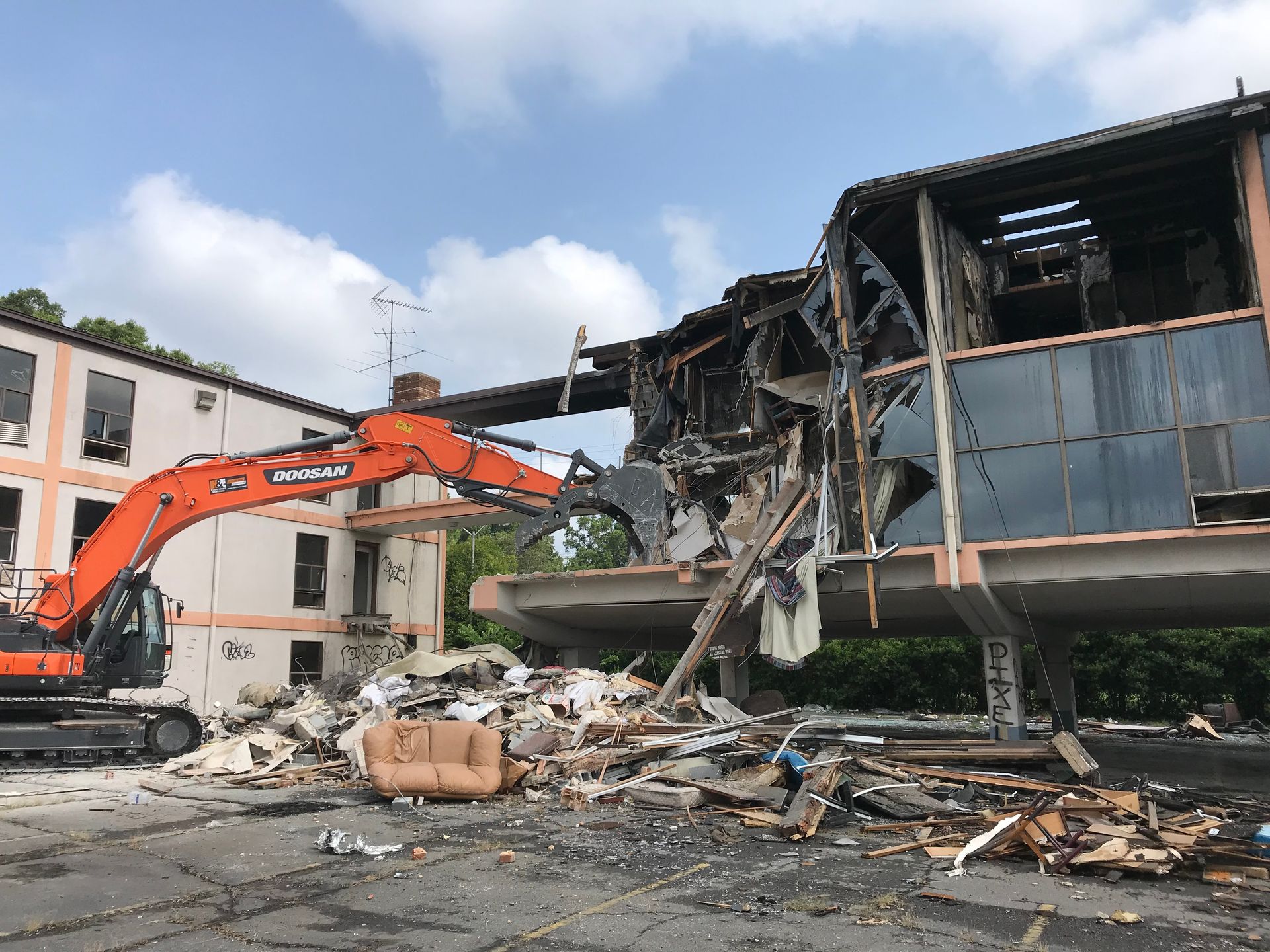 Orange excavator demolishing a damaged building amid rubble and debris under a blue sky