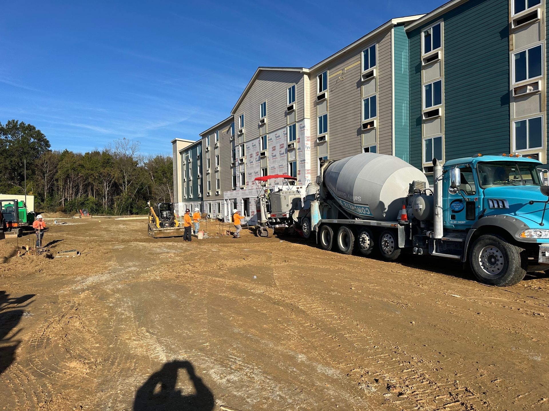 Construction site beside a long apartment building, with a cement truck on a dirt road under a blue sky