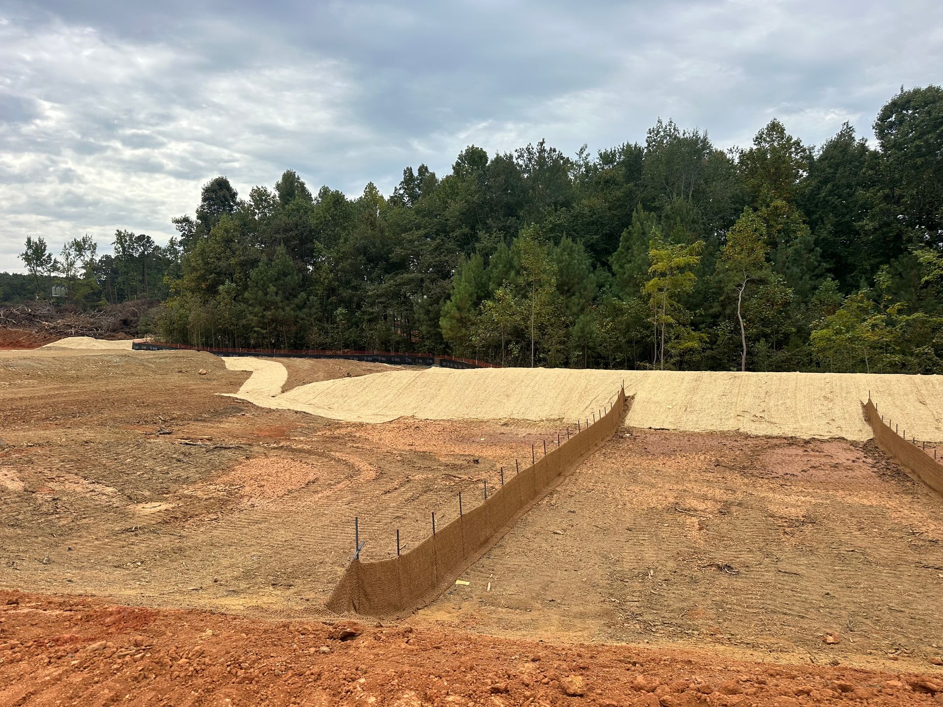 Cleared earthwork with reddish soil, pale sand, and trees under a cloudy sky