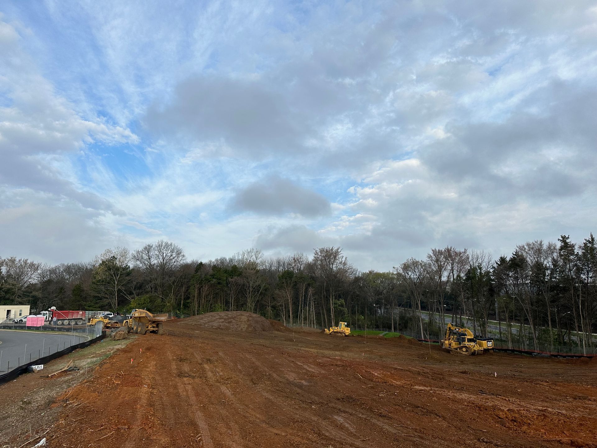Construction site with dirt lot, heavy equipment, and cloudy sky at the edge of a wooded area