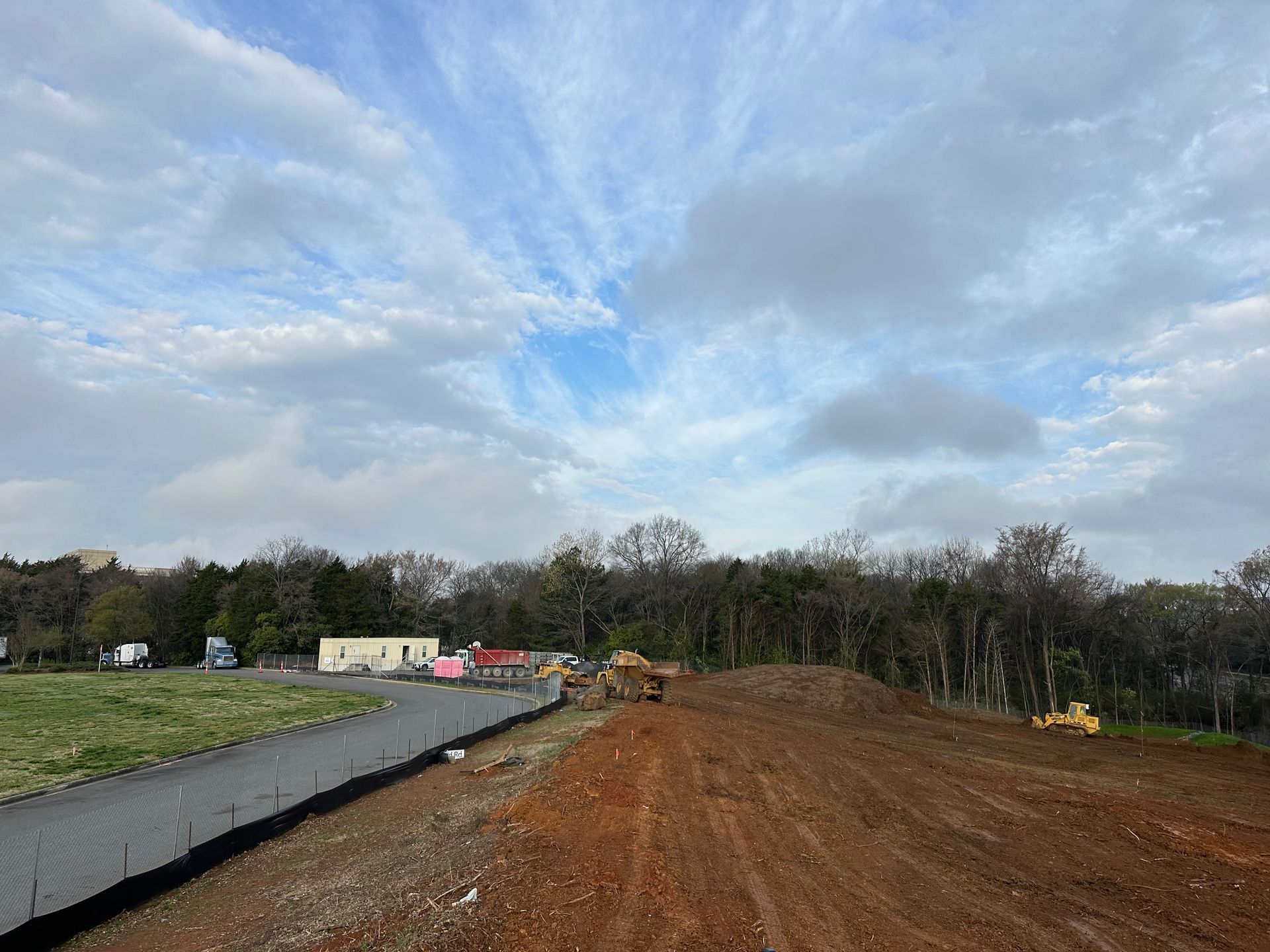 Construction site with dirt road, gravel edge, machinery, and cloudy sky over wooded background