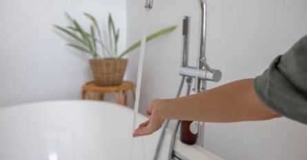 Arm reaching toward a white bathtub in a bathroom beside a chrome shower fixture and potted plant
