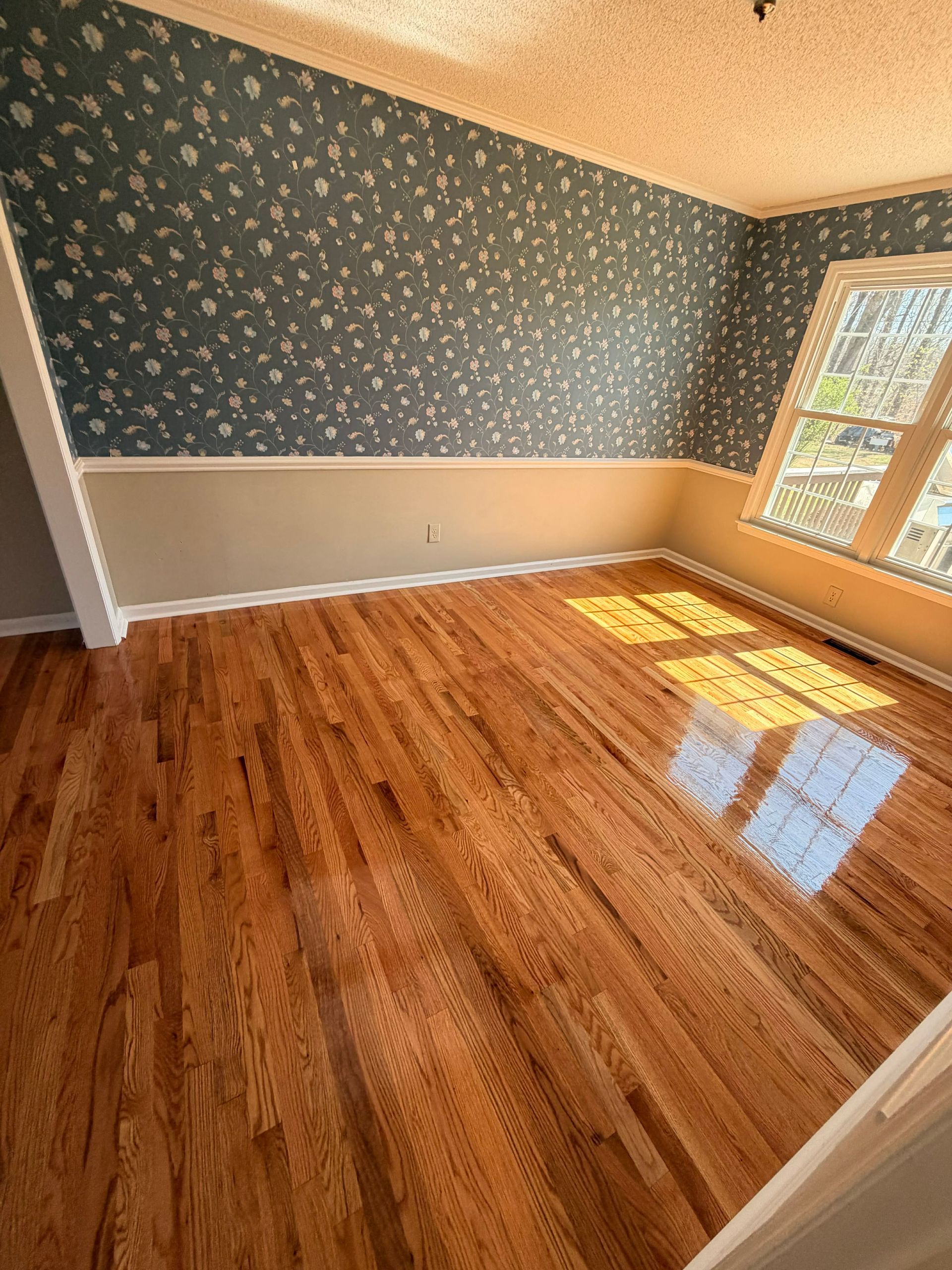 An empty room with polished hardwood floors, floral wallpaper above a tan wainscot, and sunlight streaming through a window.