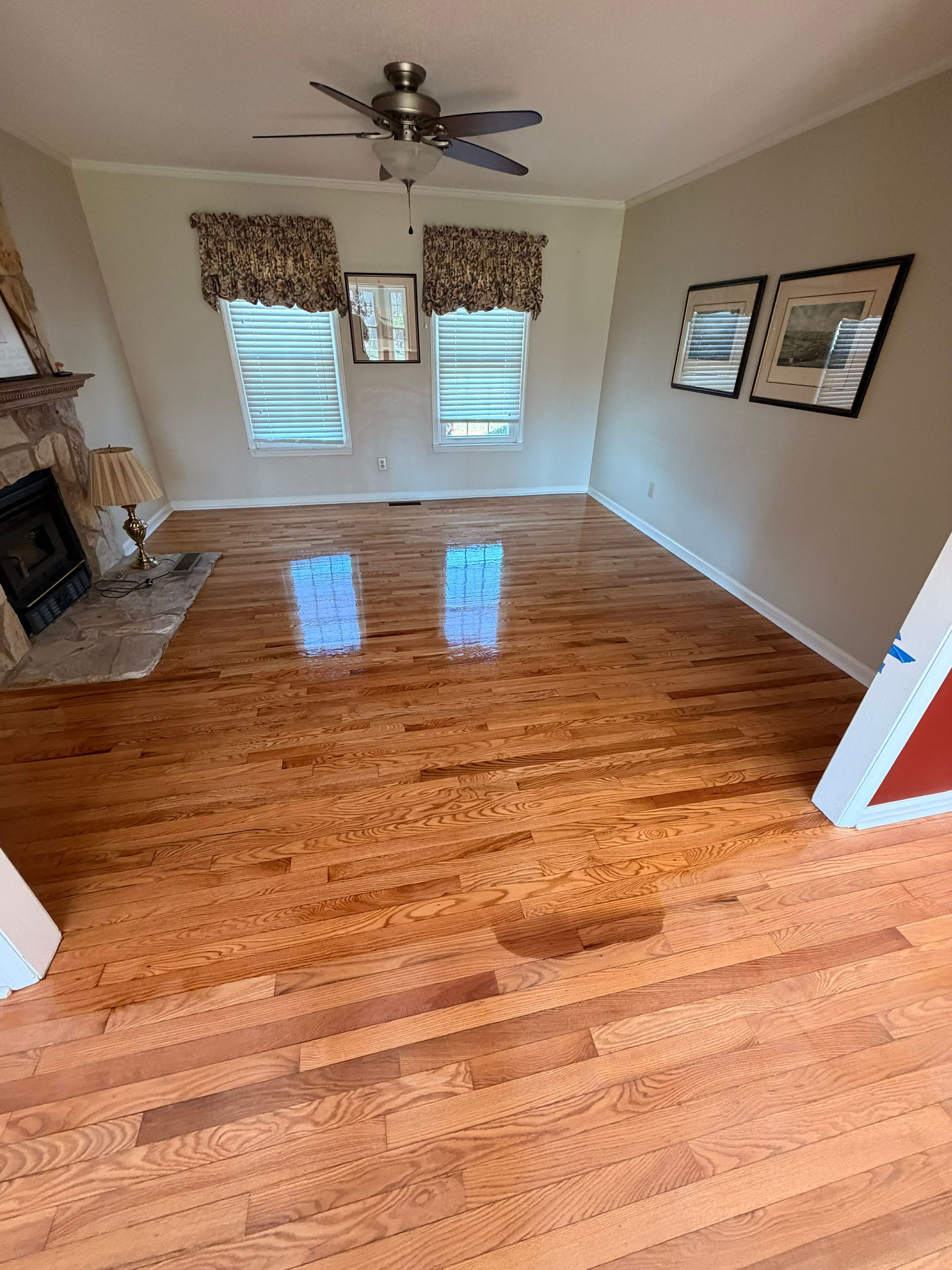 An empty room with polished hardwood floors, two windows with patterned valances, a fireplace, and wall art.