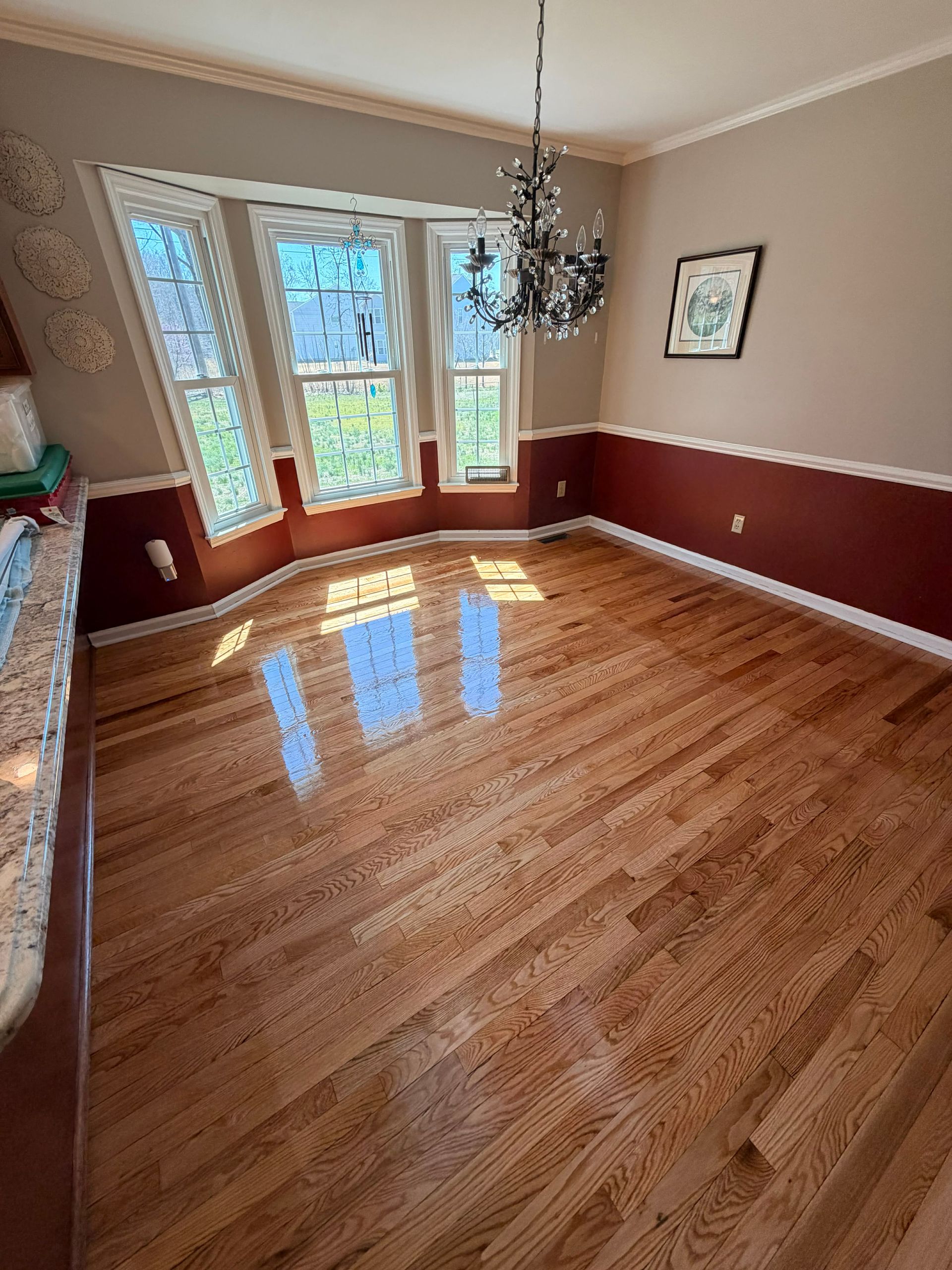 Empty dining room featuring polished hardwood floors, cream walls with burgundy wainscoting, and a crystal chandelier.