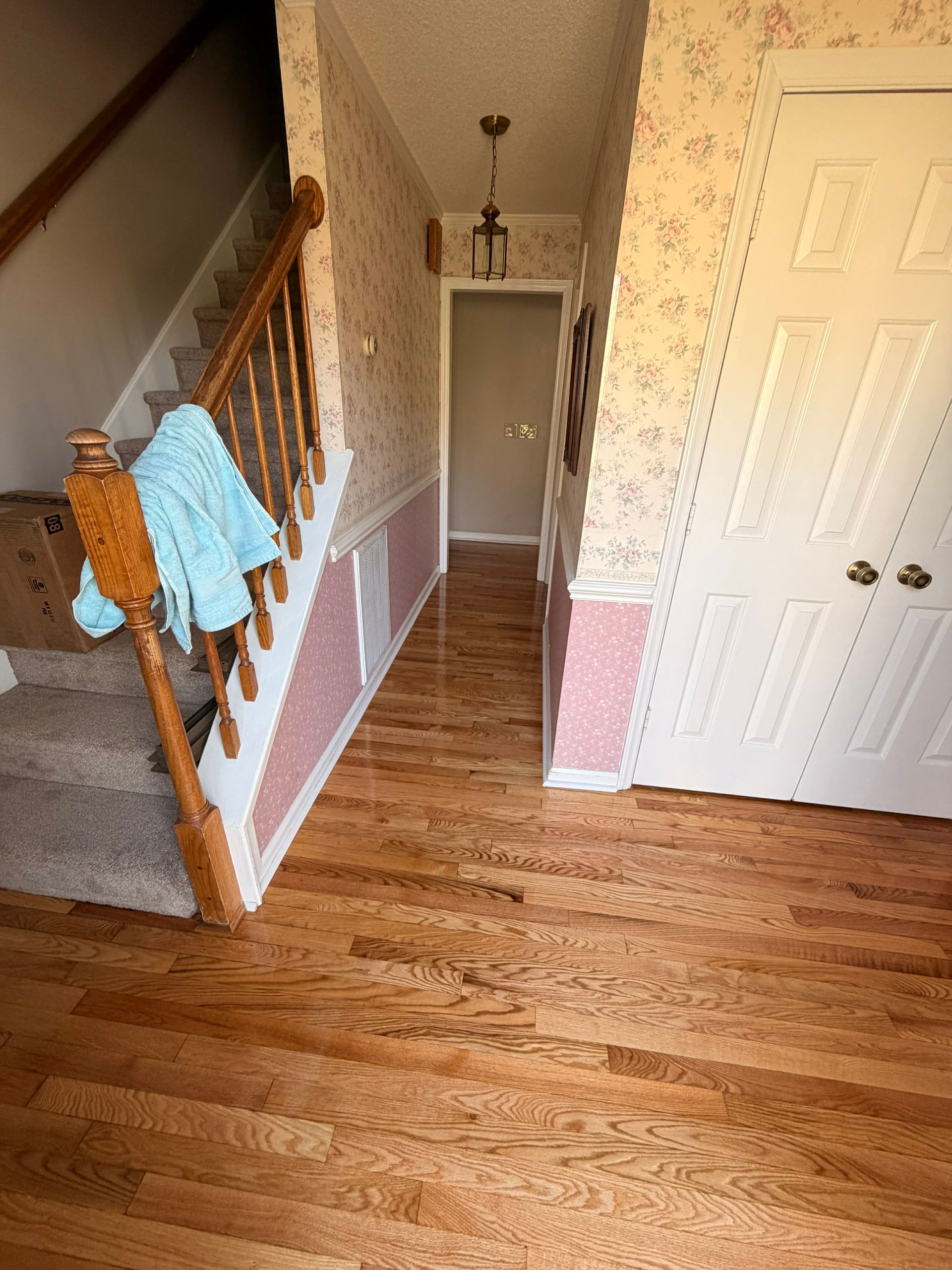 A hallway with hardwood floors, floral wallpaper, stairs with a blue towel, and white bifold doors.