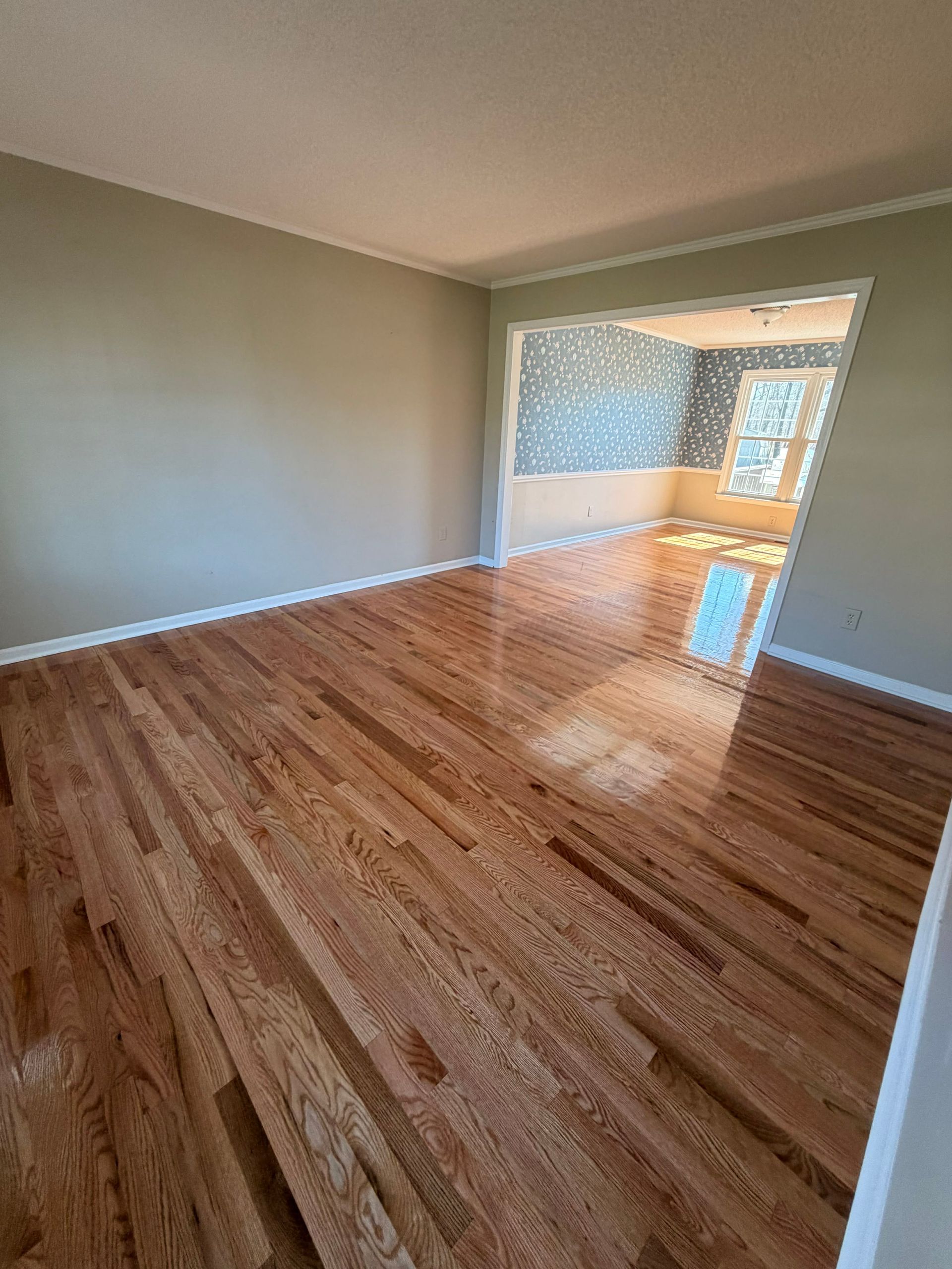 An interior view of a room with polished hardwood floors and light-colored walls, leading to an adjacent sunlit room.