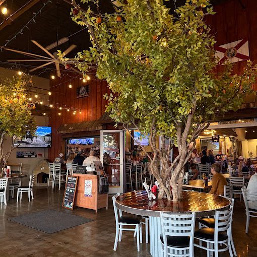 Restaurant interior with trees, tables, and customers. A bar, TVs, and a large fan are visible.