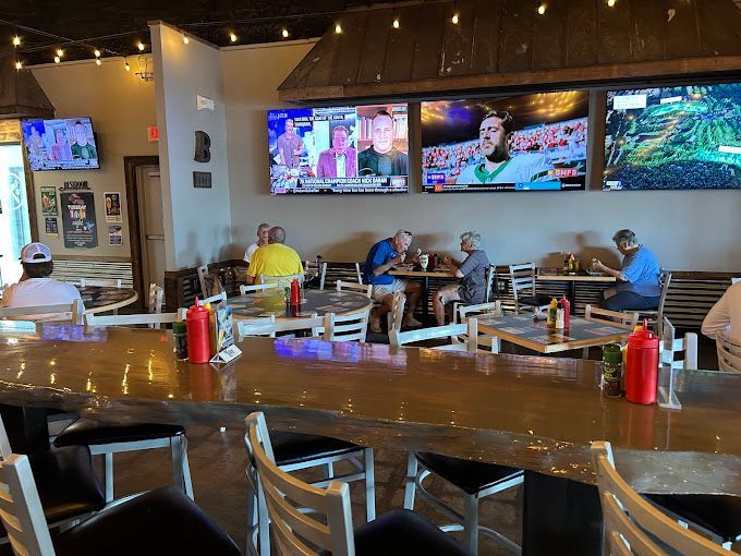 Interior of a restaurant with people seated at tables and a bar, multiple TVs mounted on the wall.