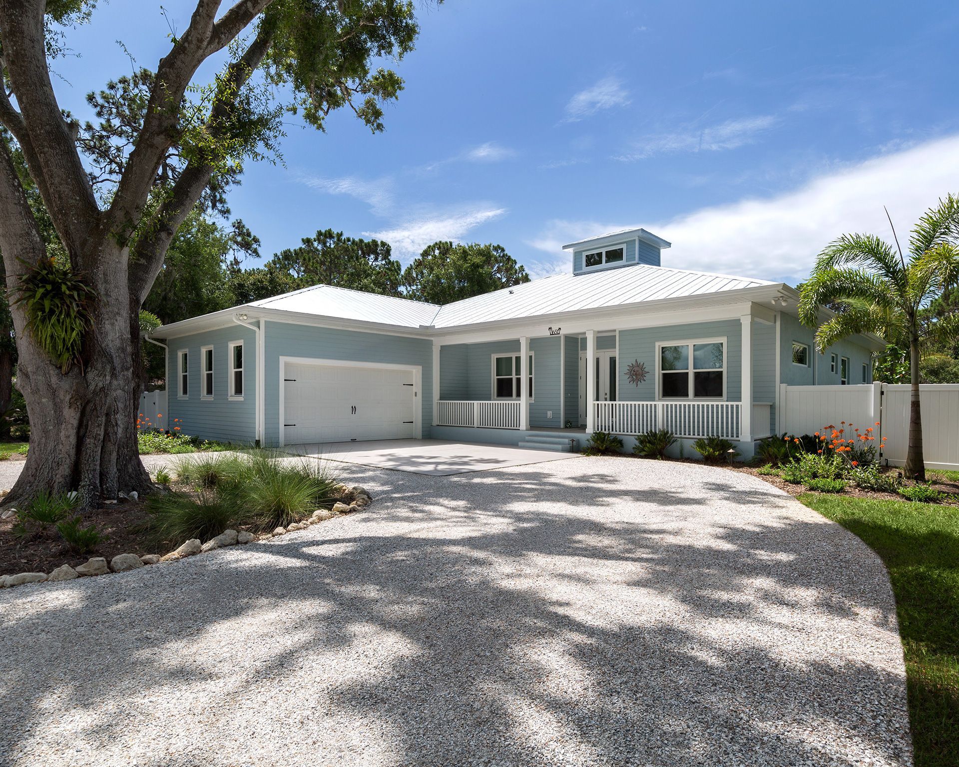 Blue coastal-style house with white trim and a gravel driveway under a sunny sky.