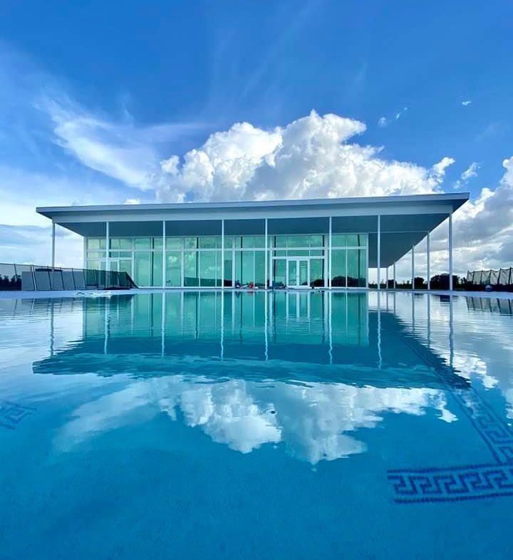 Rooftop pool with glass-walled structure reflecting the sky. Cloudy blue sky above, water reflecting clouds.