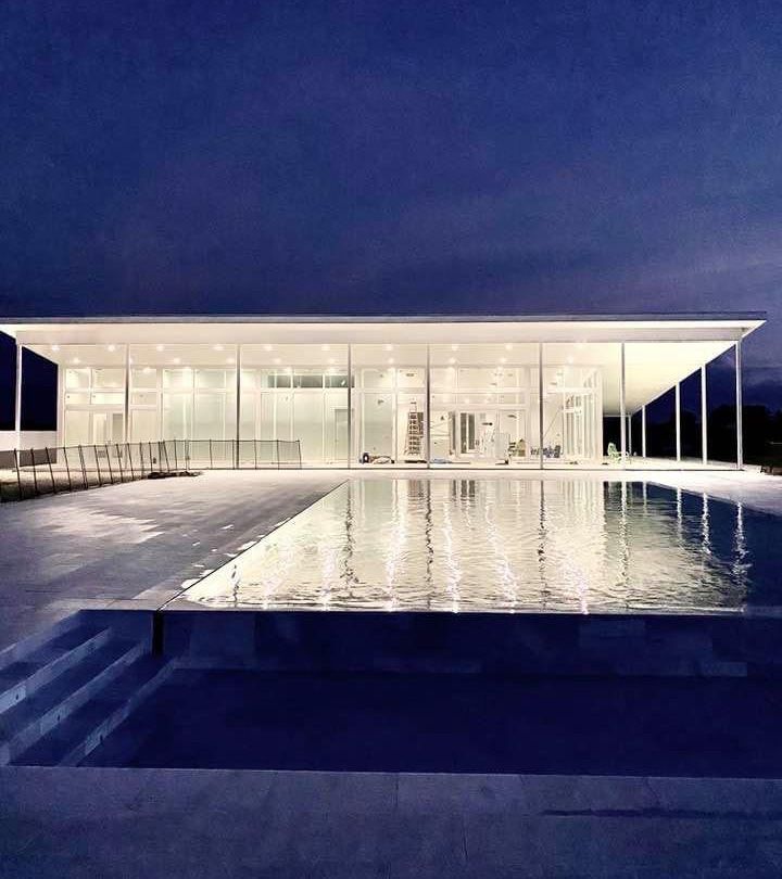 A modern glass house reflects in a pool at dusk, with a blue sky above.