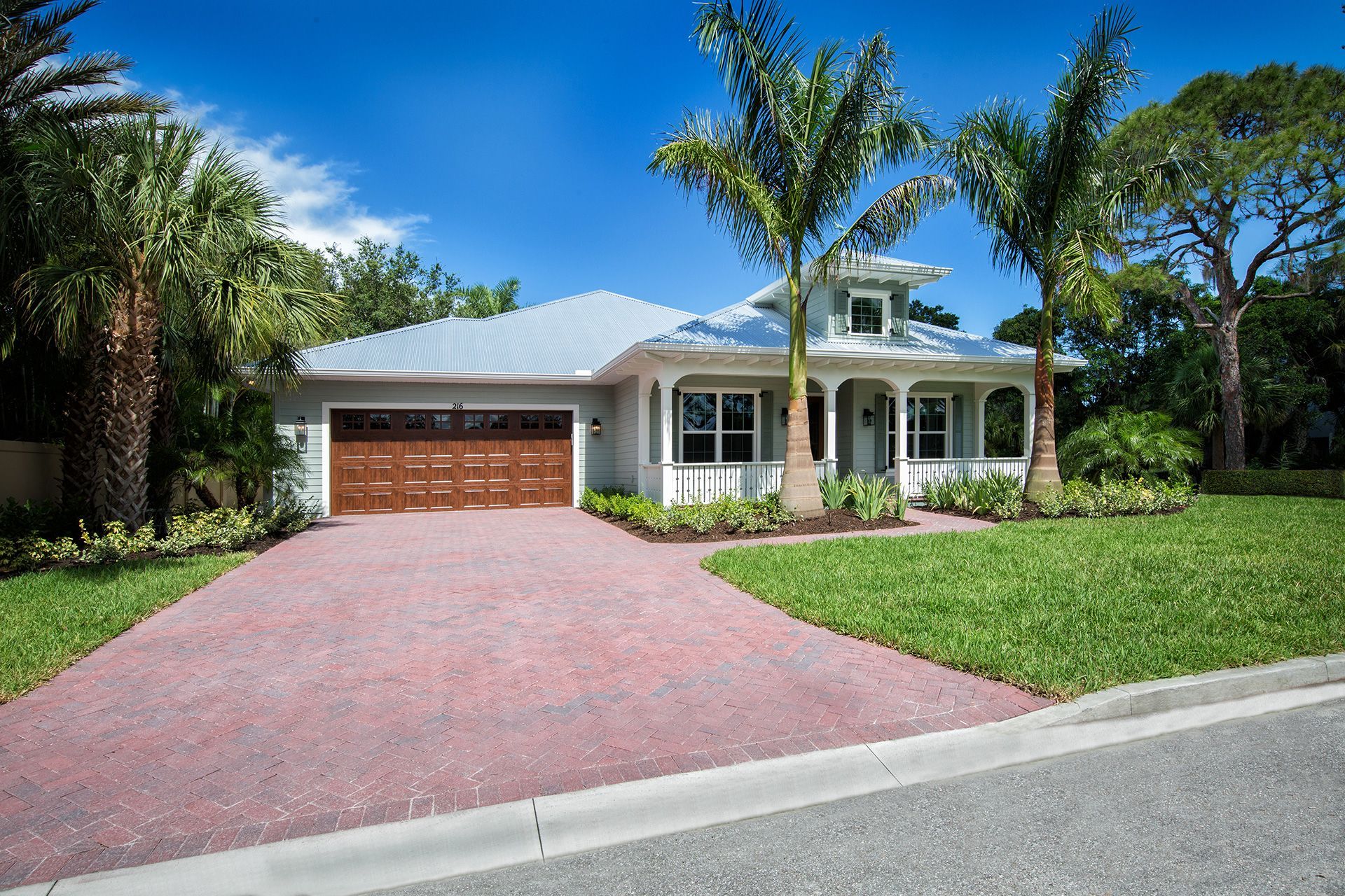 Coastal home with a red-brick driveway, a garage, and palm trees. Sunny day, blue sky.