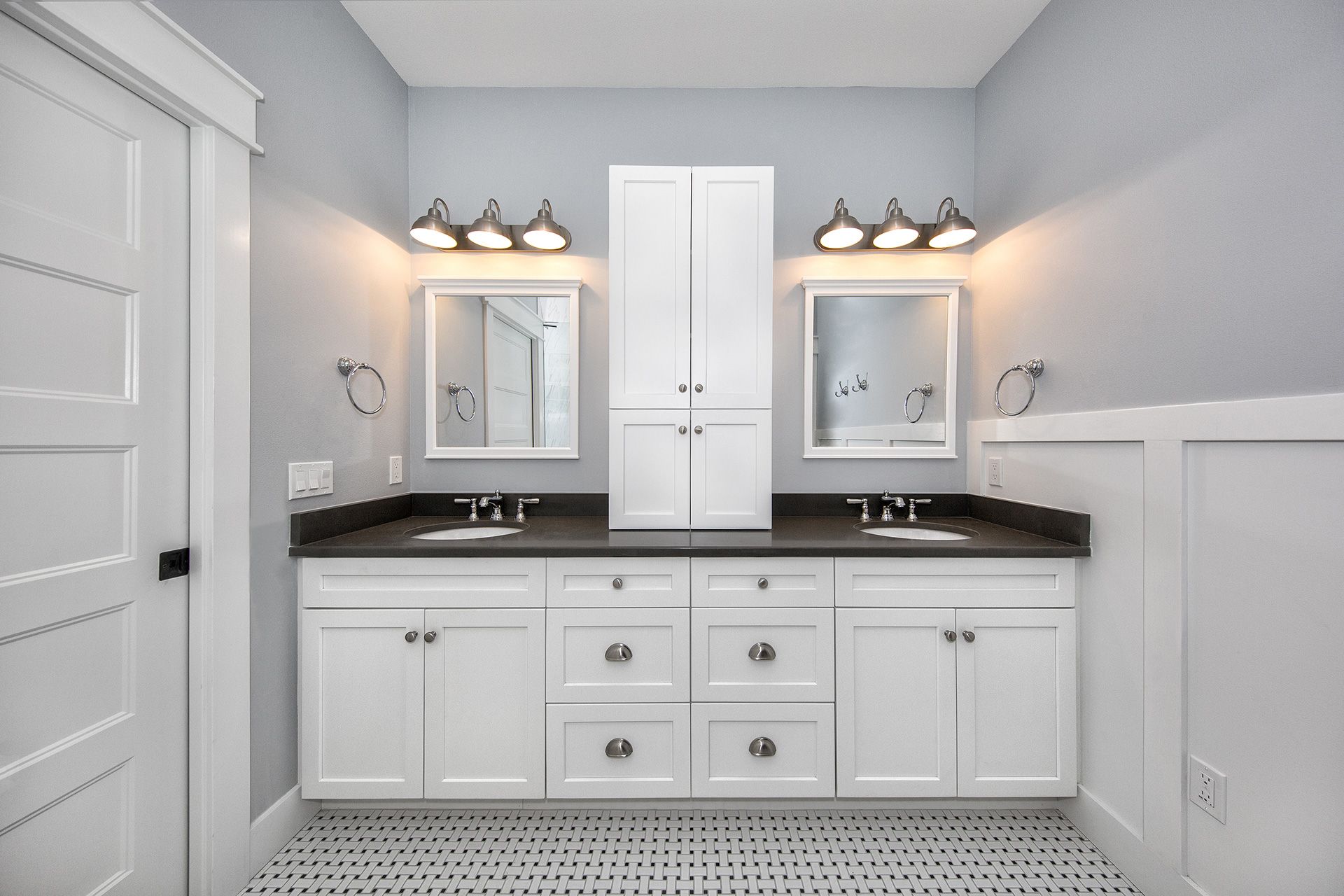 White bathroom with double sinks, mirrors, and central storage cabinet; gray walls, black countertops, and patterned floor.