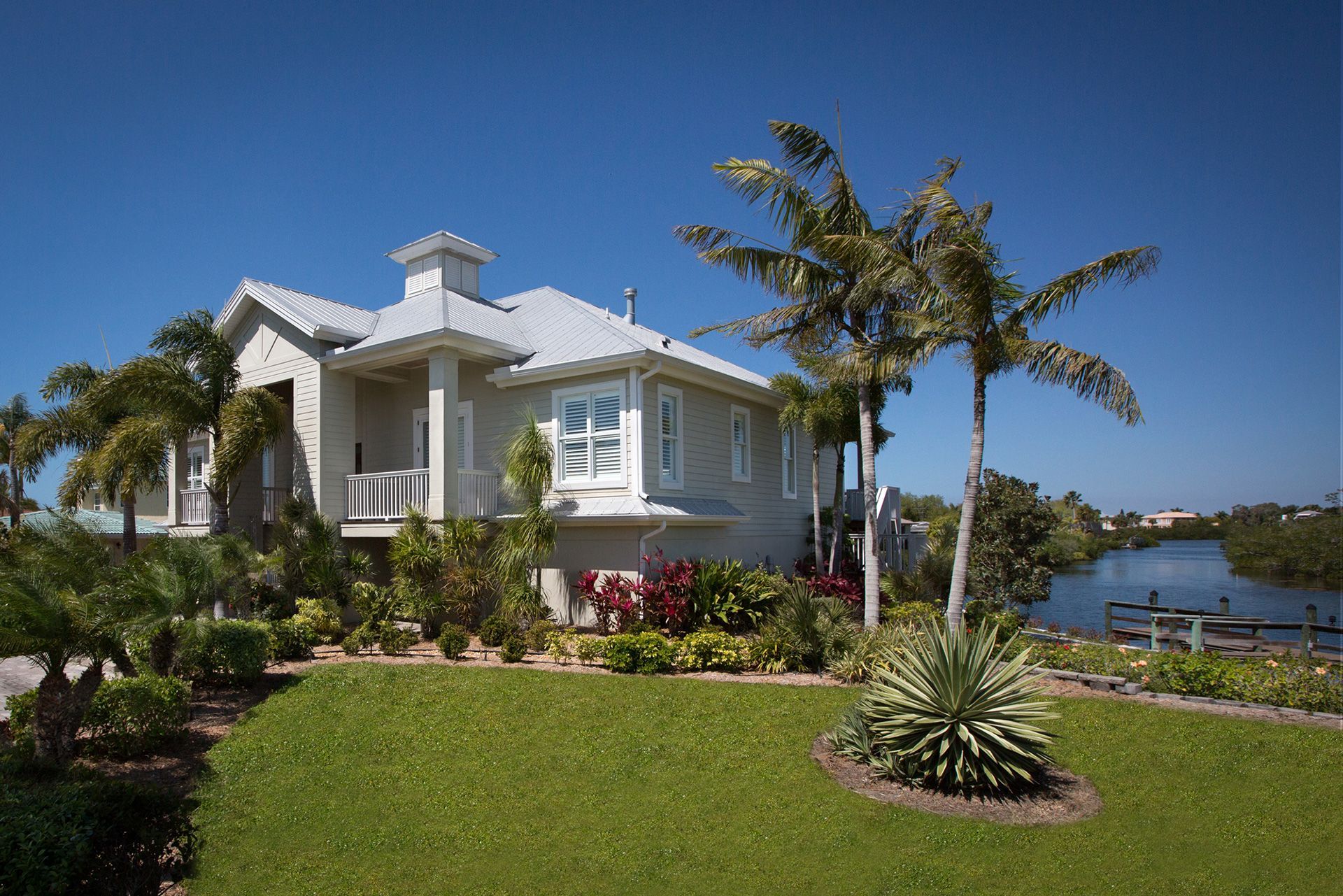 Coastal house with palm trees, green lawn, and waterway under a clear blue sky.