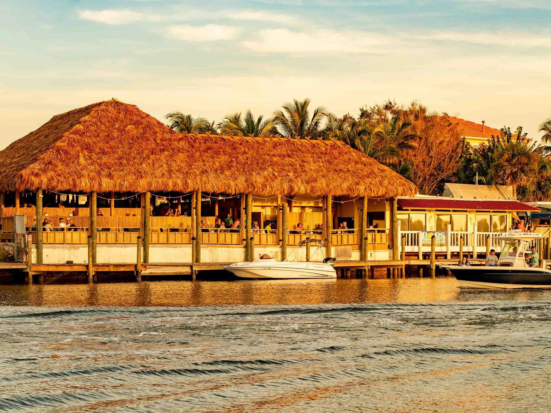 A waterfront restaurant with a thatched roof, boats docked nearby, and a sandy shoreline.