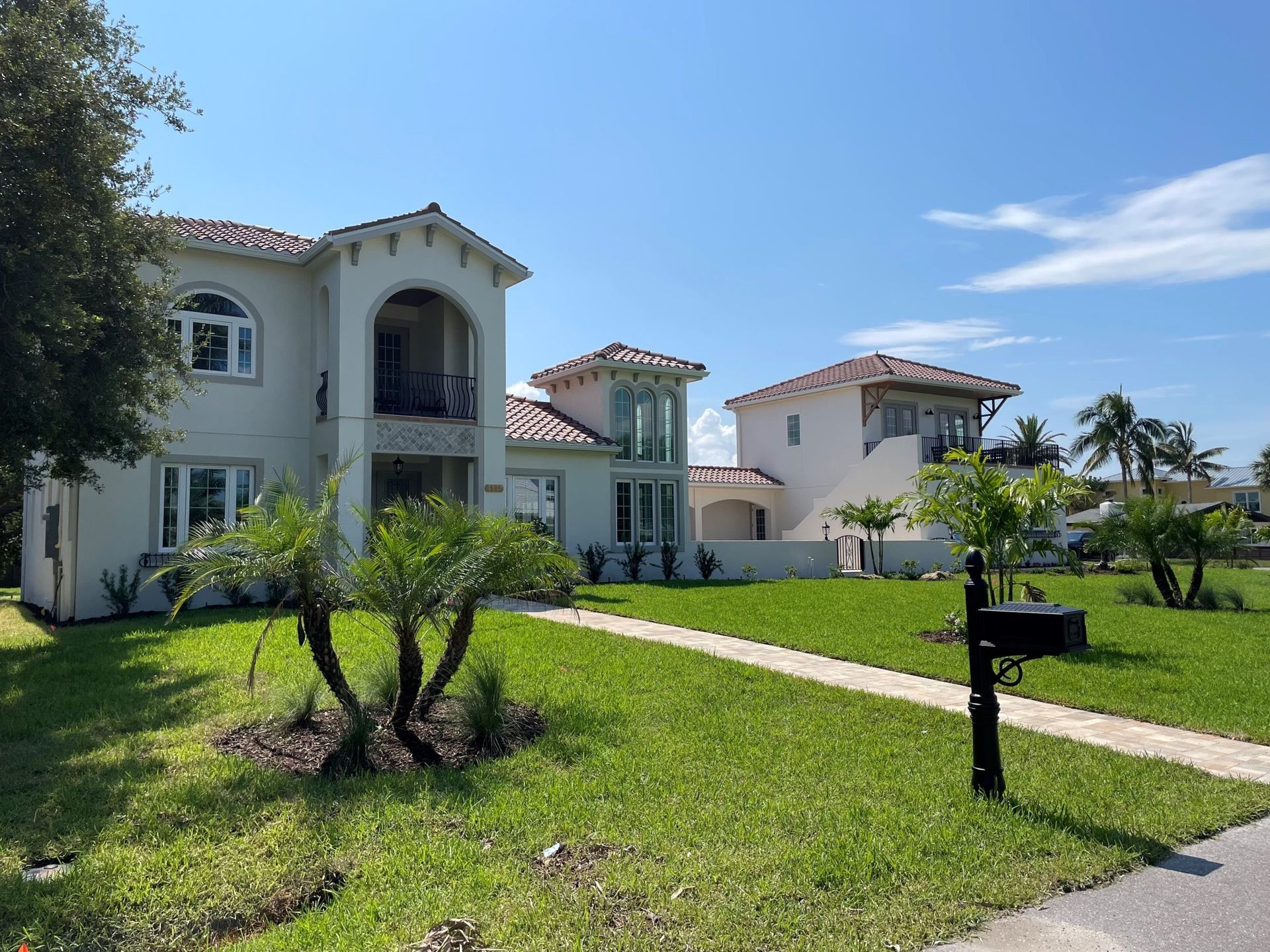 Two-story stucco houses with red tile roofs, palm trees, and green lawns under a blue sky.
