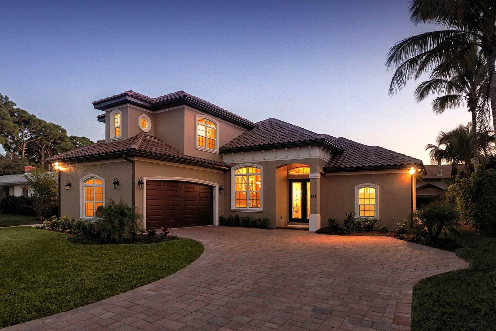 Two-story stucco home with a tiled roof, lit windows, and a brick driveway at dusk.