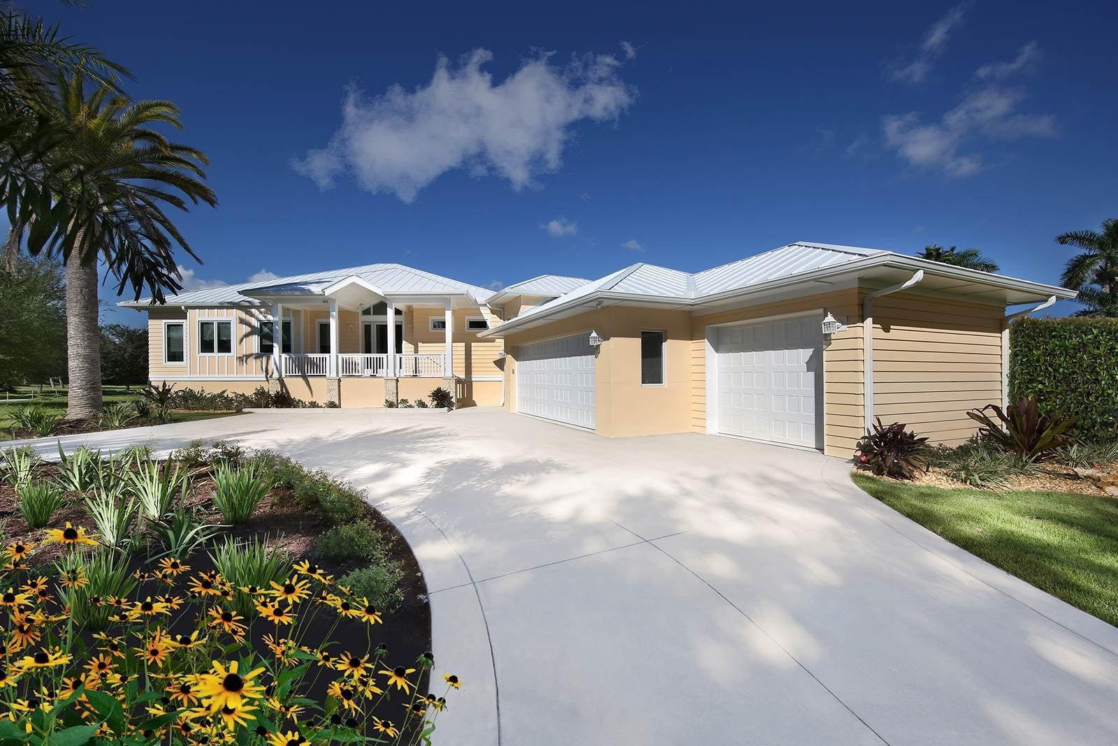 Beige house with white roof and three-car garage, long driveway, palm tree, blue sky.