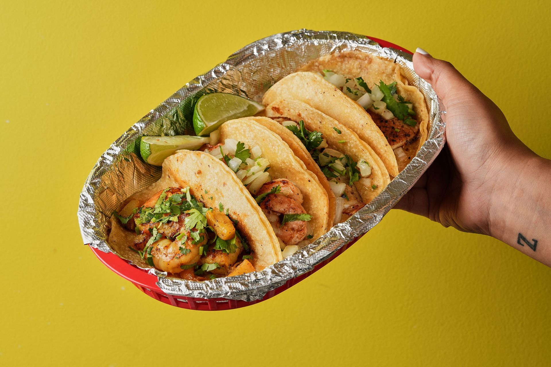 Hand holding a foil container with four tacos, garnished with cilantro and lime, on a yellow backdrop.