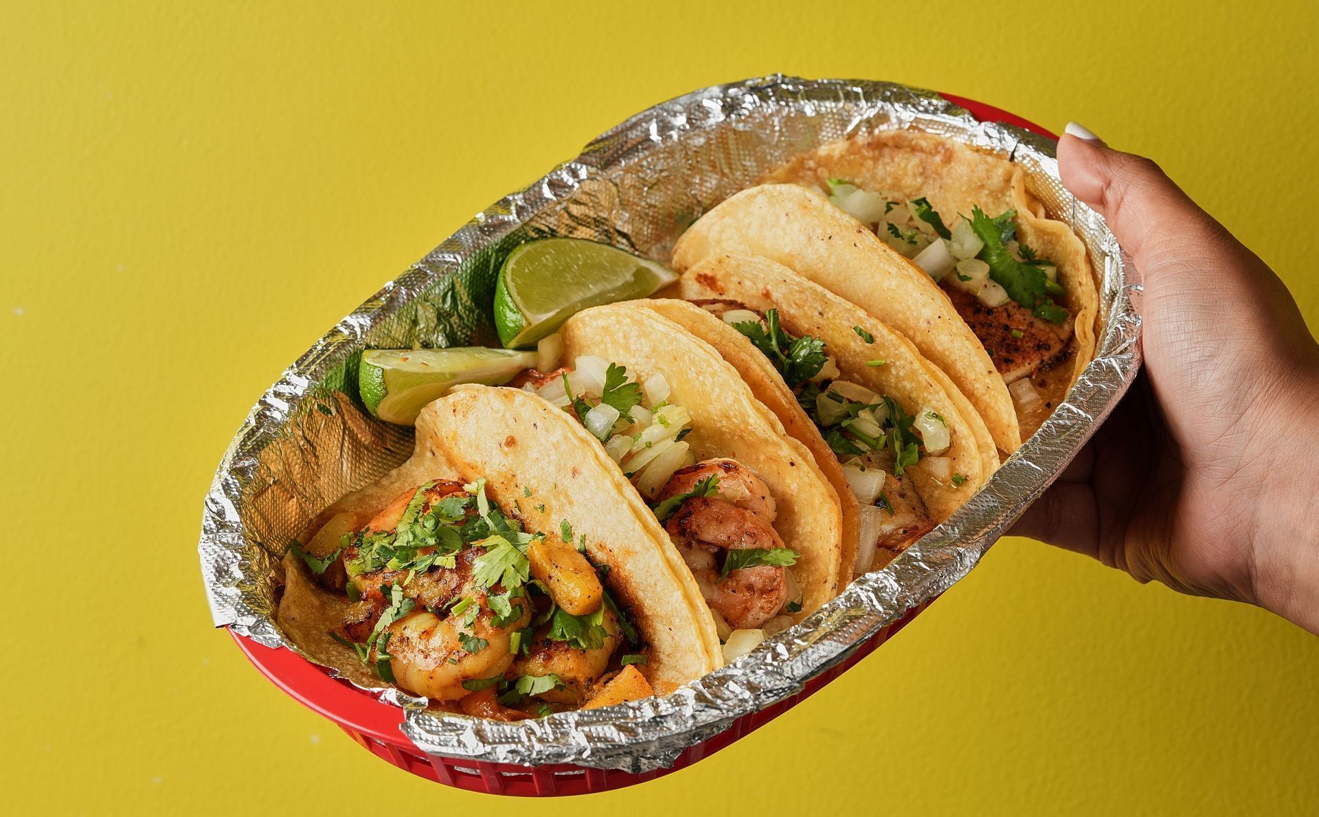 Hand holding a tray of five tacos with lime on a yellow background.