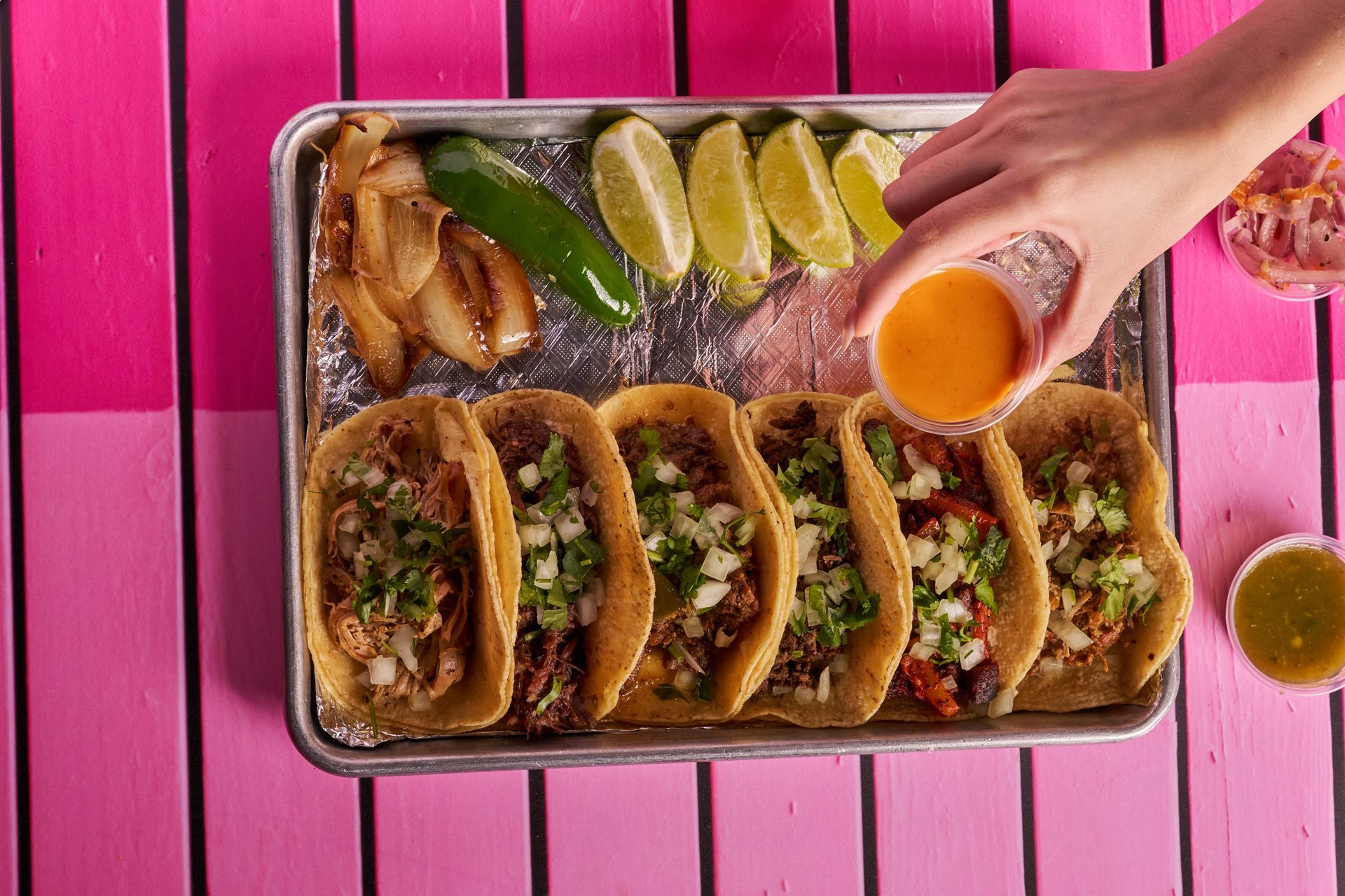 Tray of tacos with toppings and lime wedges, hand adding sauce on a pink background.